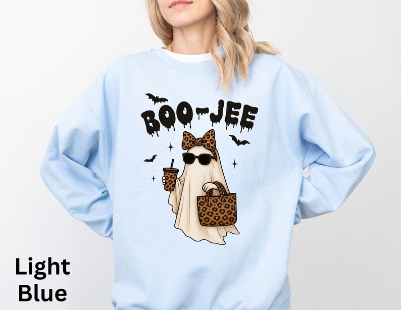 Op de afbeelding: Lichtblauwe sweatshirt met een cartoongeest met een zonnebril, een luipaardstrik en een handtas en koffiebeker met luipaardprint. De woorden "BOO-JEE" staan boven de geest. Zwarte vleermuizen en sterren maken het ontwerp compleet.