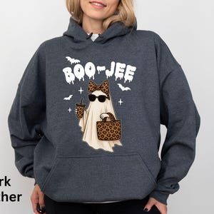 Op de afbeelding: Donkergrijze hoodie met een Halloween-thema. De afbeelding toont een spook met een zonnebril en een luipaardprint strik, met een koffiebeker en een handtas. Het woord "BOO-JEE" staat boven het spook.