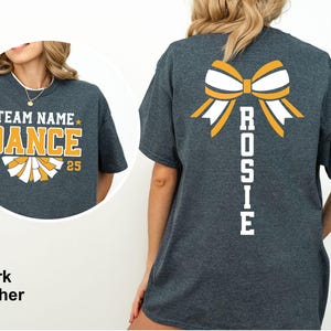 May include: Dark heather gray t-shirt with a cheerleading design. The front features the text "TEAM NAME DANCE 2025" in yellow and white. The back displays a bow graphic and the name "ROSIE" in white. A perfect tee for cheerleading.