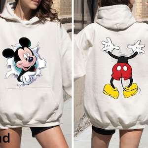 May include: Sand-colored hoodie featuring a Mickey Mouse graphic. The front shows Mickey breaking through a torn paper effect, while the back displays a full-body cartoon image of Mickey Mouse. The hoodie has a front pocket and a hood.