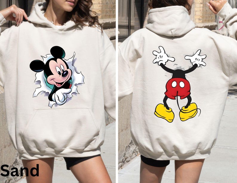 May include: Sand-colored hoodie featuring a Mickey Mouse graphic. The front shows Mickey breaking through a torn paper effect, while the back displays a full-body cartoon image of Mickey Mouse. The hoodie has a front pocket and a hood.