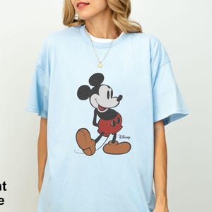 May include: Light blue t-shirt featuring a classic Mickey Mouse graphic. The cartoon character is depicted in his iconic pose, wearing red shorts, brown shoes, and black gloves. The word "Disney" is printed below the character.