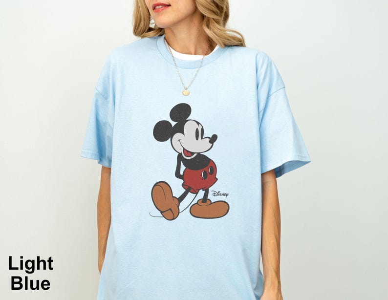 May include: Light blue t-shirt featuring a classic Mickey Mouse graphic. The cartoon character is depicted in his iconic pose, wearing red shorts, brown shoes, and black gloves. The word "Disney" is printed below the character.