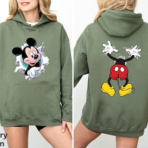 May include: Military green hoodie featuring a Mickey Mouse graphic on the front and back. The front graphic shows Mickey breaking through a torn paper effect. The back graphic shows Mickey's arms and legs. The hoodie has a kangaroo pocket.