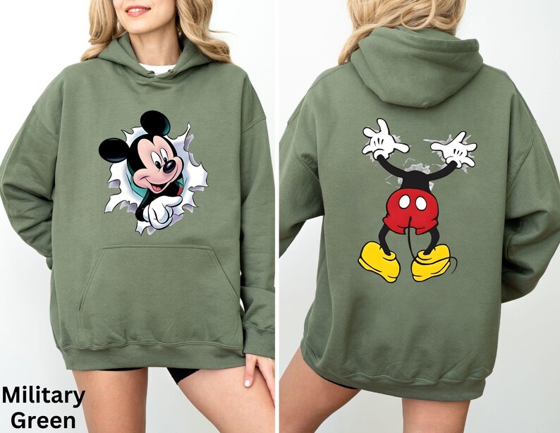 May include: Military green hoodie featuring a Mickey Mouse graphic on the front and back. The front graphic shows Mickey breaking through a torn paper effect. The back graphic shows Mickey's arms and legs. The hoodie has a kangaroo pocket.