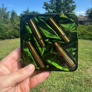 May include: A square resin coaster featuring a deep emerald green glitter background with a fabric-like texture. Four metallic brass-colored bullet casings are embedded within the resin, arranged in a diagonal pattern across the surface.