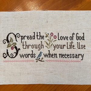 May include: Cross-stitched fabric with the text "Spread the Love of God through your Life. Use words when necessary." The text is in black, with decorative floral and bird accents. The fabric is a light beige color.