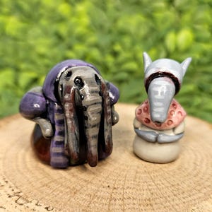 Könnte beinhalten: Zwei skurrile Keramikfiguren von elefantenartigen Kreaturen. Eine trägt einen lila Kapuzenmantel, die andere einen grauen Hut und einen rosa Schal. Sie stehen auf einer Holzoberfläche vor einem verschwommenen grünen Hintergrund.