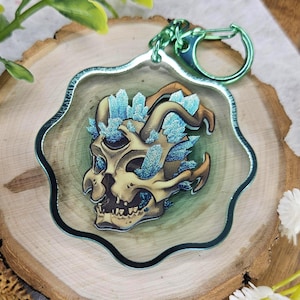 May include: A clear acrylic keychain featuring a beige skull with three eye sockets and curved horns, adorned with sparkling blue crystal clusters. The keychain has a scalloped edge and a metallic green lobster clasp attached at the top.