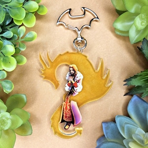 Könnte beinhalten: Ein goldfarbener Drachen-Schlüsselanhänger mit einem silbernen, fledermausförmigen Verschluss. Der Schlüsselanhänger zeigt eine detaillierte Illustration einer Person in einem rot-goldenen Outfit. Der Hintergrund ist hellbraun, und es gibt grüne Pflanzen an den Rändern.