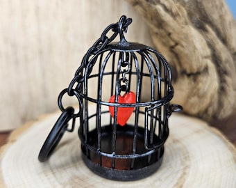 Puede incluir: Un llavero de jaula para pájaros de metal negro con un pequeño dije de corazón rojo brillante suspendido en el centro. La jaula tiene una parte superior redondeada con una argolla de metal resistente y una cadena, mostrando una estructura de alambre.