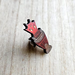 Puede incluir: Un pin de madera con una criatura roja estilizada con una cabeza en forma de corona y patrones geométricos. El pin tiene un aspecto texturizado con acentos marrones y grises, y un respaldo de alfiler para la fijación.