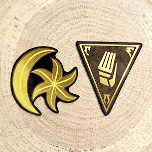 Puede incluir: Dos parches bordados. Uno presenta un diseño de media luna dorada y estrella sobre un fondo negro. El otro es un triángulo marrón con una mano y un símbolo dorados. Ambos están sobre una superficie de vetas de madera.