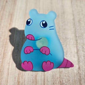 May include: A light blue plastic figurine of a cartoon hamster with pink feet and a light green nose. The hamster has black eyes and whiskers.
