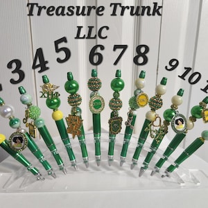 May include: Twelve green and gold pens with various charms and beads. The pens are displayed on a clear plastic stand. The numbers 1 through 12 are displayed above the pens.