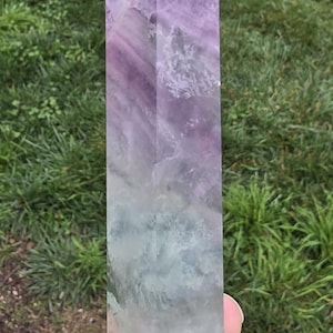 May include: A tall, rectangular fluorite crystal point. The crystal is a gradient of colors, transitioning from a purple tip to a green base. The stone is held against a backdrop of green grass.