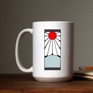 May include: White ceramic mug with a black and white graphic of a sun with a red center and a white background. The graphic is surrounded by a black border.