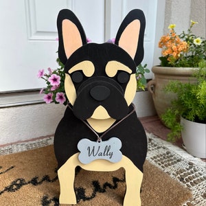 May include: A wooden French Bulldog cutout painted in black and cream with large upright ears. The dog figure wears a gray bone-shaped tag hanging from a string around its neck, with the name Wally engraved in a dark script font on the tag.