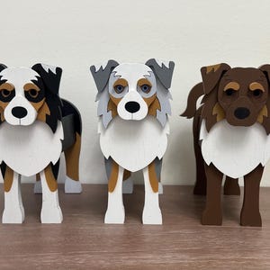 May include: Three wooden dog sculptures in various colors. The dogs have white chests and legs, with different colored heads and ears. One dog is black, brown, and white, another is gray and white, and the third is brown and white.