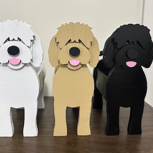 May include: Three wooden dog planter boxes, one white, one brown, and one black. Each planter has a cartoon dog face with a pink tongue.