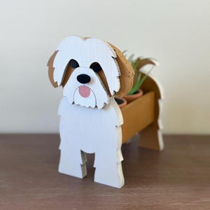 May include: A wooden dog-shaped planter featuring a white and brown painted finish. The dog has black eyes, a black nose, and a pink tongue. The body of the dog forms a hollow container designed to hold small potted plants, with visible green foliage.