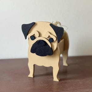 May include: A wooden pug dog figurine with a light tan body, black ears, and a black snout. The pug has a sad expression with round black eyes. The figurine is standing on four legs and appears to be a decorative item.