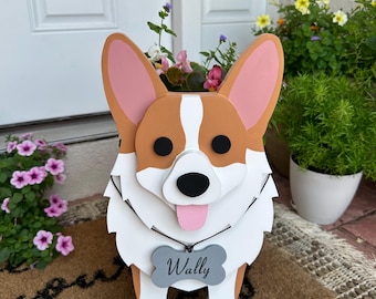 May include: A wooden planter shaped like a corgi dog with a white chest and a pink tongue sticking out. The planter has a gray bone-shaped tag with the name "Wally" written on it. The planter is sitting on a brown doormat.