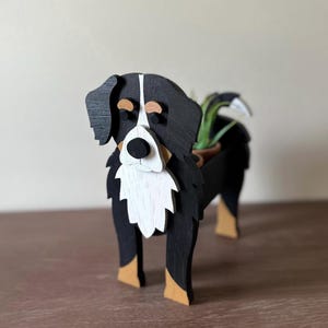 May include: A wooden Bernese Mountain Dog planter, painted black, white, and tan. The dog-shaped planter holds a small potted plant. The dog has a white chest, muzzle, and a black nose. The ears and body are black.