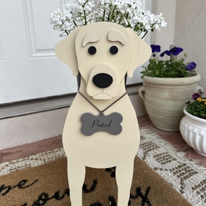 May include: A wooden planter shaped like a yellow Labrador Retriever with black eyes and nose. The planter has a gray bone-shaped tag that says "Pretzel". The planter is filled with white flowers.
