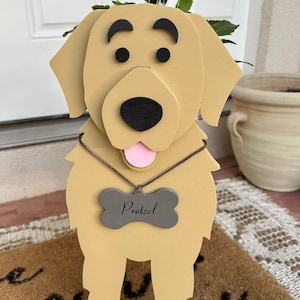 May include: A wooden planter shaped like a golden retriever dog with a pink tongue sticking out. The dog is wearing a brown rope necklace with a gray bone tag that says "Pretzel". The planter is holding purple and white flowers.