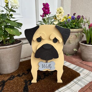 May include: A wooden pug-shaped planter with black ears and muzzle, holding a light blue tag that reads "Wally." The planter is filled with vibrant purple and yellow flowers. It sits on a brown doormat, with other potted plants in the background.