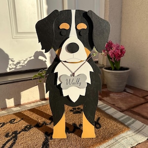 May include: A wooden dog-shaped planter with black, white, and tan coloring. The dog has a tag that says "Wally". The planter is standing on a brown doormat with the words "wipe your paws" on it.