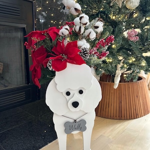 May include: White wooden poodle cutout with a gray bone-shaped tag that says "Wally". The poodle is decorated with red poinsettia flowers and white cotton bolls. The poodle is standing in front of a Christmas tree.