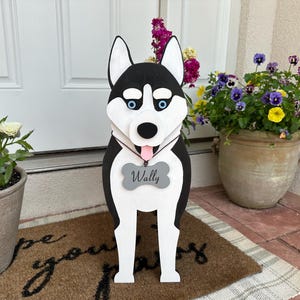 May include: A wooden husky dog decoration with black, white, and blue details. The dog has a gray bone-shaped tag that says "Wally". The dog is standing on a brown mat in front of a white door, with potted plants on either side.