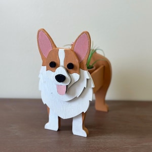 May include: A wooden planter shaped like a corgi dog. The planter is painted white with brown and pink accents. The dog has black eyes and a pink tongue sticking out. The planter has a small pot for a plant in the back.