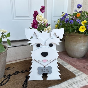 May include: A white wooden dog-shaped planter with black eyes and nose, and a pink tongue. A gray bone-shaped tag reads "Wally". The planter is filled with colorful flowers and sits on a welcome mat.