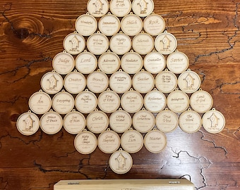 Può includere: Set di ornamenti per albero di Natale in legno con vari titoli religiosi e una scatola di legno. Gli ornamenti circolari presentano una figura ritagliata e testo. La scatola rettangolare ha un'immagine incisa e testo.