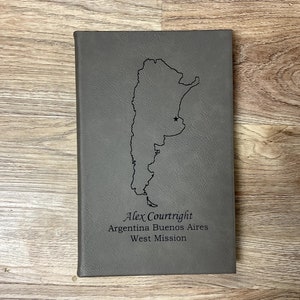 May include: Gray journal with a debossed outline of Argentina and a star marking a location. The text "Alex Courtright, Argentina Buenos Aires, West Mission" is below the map. The journal is on a wooden surface.