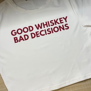 May include: White cropped t-shirt with the text "GOOD WHISKEY BAD DECISIONS" printed in red.