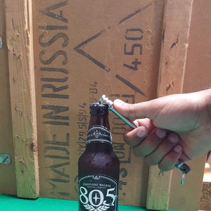 Könnte beinhalten: Ein Satz von sieben silbernen Flaschenöffnern mit Schlüsselringen. Die Flaschenöffner sind aus Metall und haben ein schlichtes Design. Die Flaschenöffner sind fächerförmig auf einer grünen Oberfläche angeordnet. Im Hintergrund befindet sich eine braune Flasche Firestone Walker 805 Brewing Company Bier.