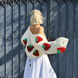 May include: A white knitted jumper with red and green strawberry appliques. The jumper is worn with a white skirt and brown boots.