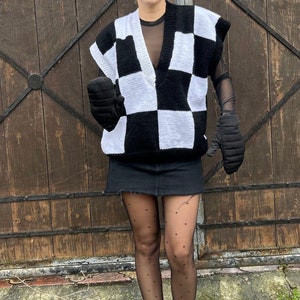 Peut inclure: Un gilet en tricot à carreaux noir et blanc avec un col en V. Le gilet est porté sur une chemise noire et associé à une mini-jupe noire et des collants noirs à pois. La personne porte des baskets montantes noires.