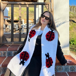 May include: White knitted scarf with red and black ladybugs. The scarf is worn by a person wearing a black jacket and sunglasses.