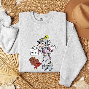Peut inclure: Un sweat-shirt gris clair avec un personnage de dessin anim&eacute; en combinaison spatiale tenant une pancarte avec l'inscription "TO MARS". Le personnage est &agrave; c&ocirc;t&eacute; d'une valise marron et d'une &eacute;charpe rouge. Le sweat-shirt a des manches longues.