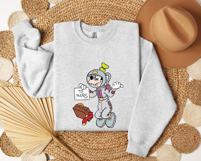 Peut inclure: Un sweat-shirt gris clair avec un personnage de dessin anim&eacute; en combinaison spatiale tenant une pancarte avec l'inscription "TO MARS". Le personnage est &agrave; c&ocirc;t&eacute; d'une valise marron et d'une &eacute;charpe rouge. Le sweat-shirt a des manches longues.