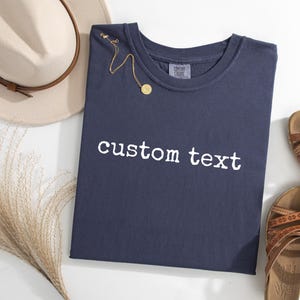 May include: A navy blue crew neck t-shirt with the white text custom text printed on the front. The shirt is displayed alongside a beige fedora hat, a gold chain necklace with a small circular pendant, and a pair of brown strappy sandals.