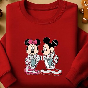 Peut inclure: Un sweat-shirt rouge &agrave; col rond avec un dessin de Mickey et Minnie Mouse en combinaisons d'astronautes. Minnie porte un n&oelig;ud rose et le nom "Sophia" est &eacute;crit sur son oreille. L'oreille de Mickey dit "Noah". Les deux personnages tiennent des casques.