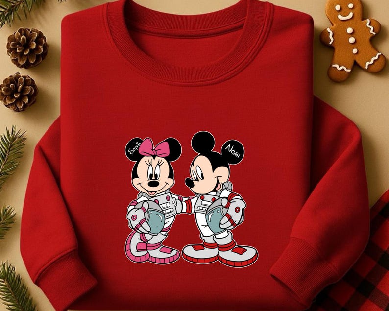 Peut inclure: Un sweat-shirt rouge &agrave; col rond avec un dessin de Mickey et Minnie Mouse en combinaisons d'astronautes. Minnie porte un n&oelig;ud rose et le nom "Sophia" est &eacute;crit sur son oreille. L'oreille de Mickey dit "Noah". Les deux personnages tiennent des casques.