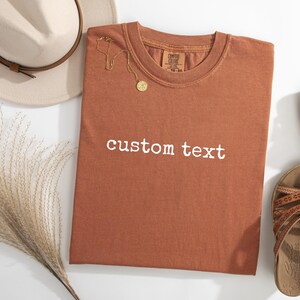 May include: A rust-colored short-sleeve t-shirt with the words custom text printed in white across the chest. The shirt is displayed alongside a beige fedora hat, a gold-toned necklace with a circular pendant, and a pair of brown strappy sandals.