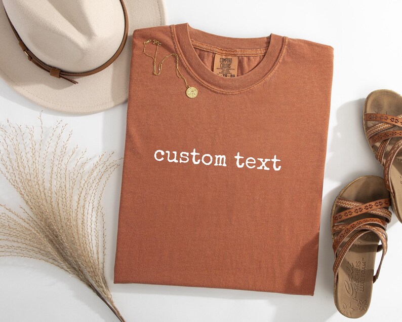 May include: A rust-colored short-sleeve t-shirt with the words custom text printed in white across the chest. The shirt is displayed alongside a beige fedora hat, a gold-toned necklace with a circular pendant, and a pair of brown strappy sandals.